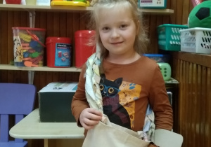 Dziewczynka pokazuje torbę przewieszoną przez ramię. Jest kremowa a na środku wyszyte serce.